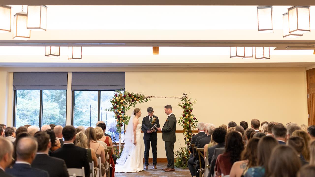 Indoor wedding ceremony with the bride and groom standing under a floral arch, surrounded by family and friends in a bright, spacious room.