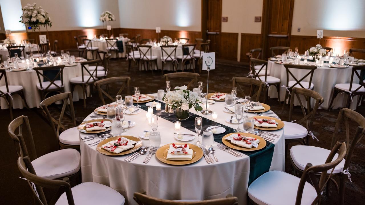 Banquet hall arranged with round tables, white linens, floral centerpieces, and golden chargers, ready for a formal wedding reception.