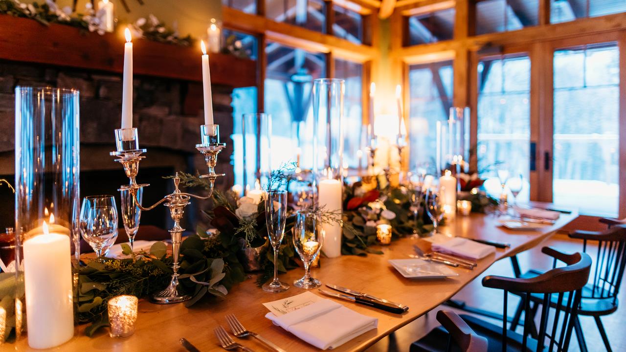 Elegant dinner table setup with lit candles, greenery, and glassware in a cozy lodge setting, featuring a warm fireplace and wooden beams.