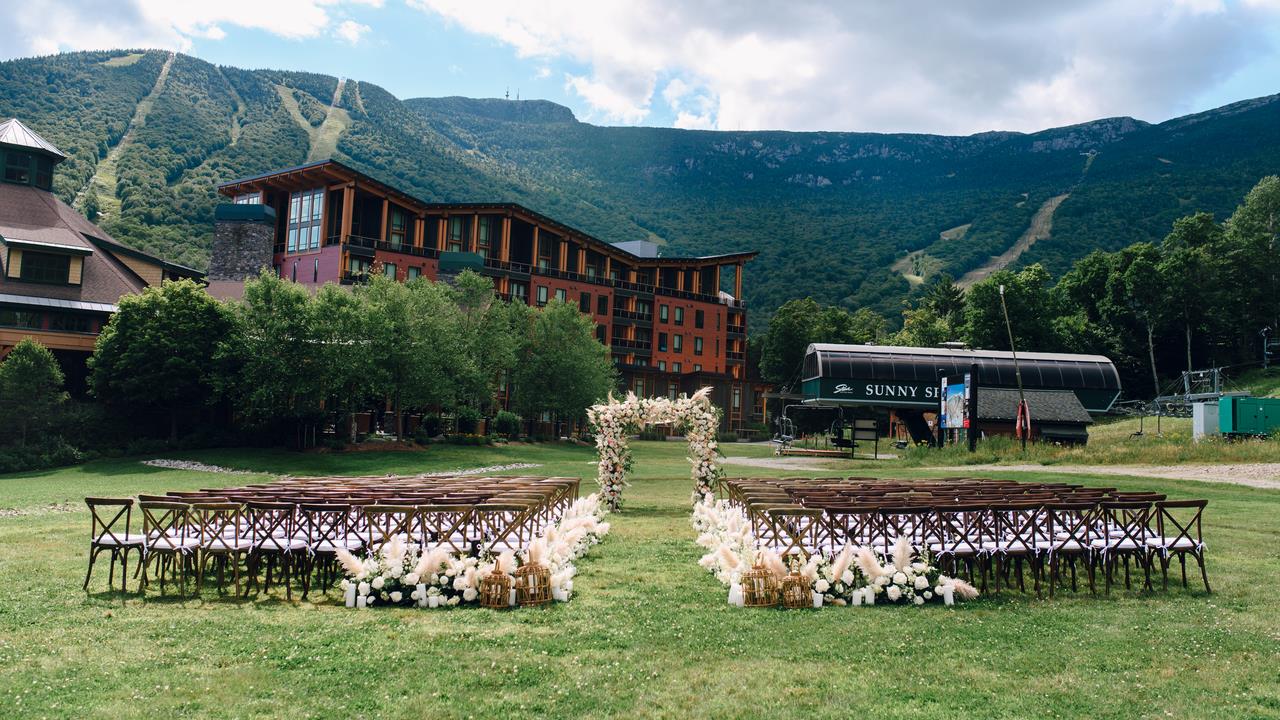 Outdoor wedding ceremony setup with wooden chairs and a floral arch, overlooking lush green mountains and a rustic lodge.